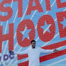 A young Black man smiles with his arms spread wide in front of a blue and white mural reading "statehood."