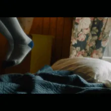 A pair of children's feet in white socks are visible jumping mid-air from a bed with a blue blanket