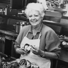 Marcella stands in the kitchen in an apron before a bunch of lettuce.