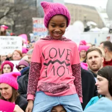 A feminine african-american child in a pink "pussyhat" on someone's shoulders during a woman's rights march. 