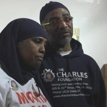 A still from "Murders That Matter": Protagonist Movita Johnson-Harrell, a middle-aged Black woman in a white t-shirt and black hijab, stands next to a middle aged Black man wearing a blue hoodie that says "The Charles Foundation"