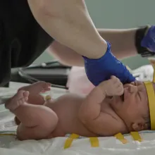 A pair of hands wearing medical gloves cares for a crying newborn infant. The nurse is using a tape measure to measure the circumference of the baby's head.
