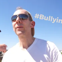 Two White men in sunglasses, one with a shirt off, stand before a green field outside on a sunny day.