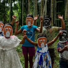 6 young children, in two rows of three, stand with their hands awkwardly raised in celebration. Behind them is a lush, green forest with tall and slim trees. The children are all wearing a paper mask that is a simple drawing of a young Hoolock Gibbon called Twiki. It is Twik's first birthday although he is not in the picture. We assume he is somewhere up in the trees behind them.