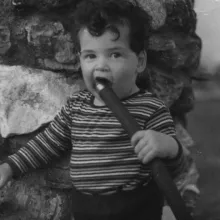 A white male toddler stands in front of a stone wall, chewing on a garden hose.