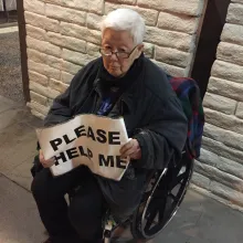 an elderly asian individual in a wheelchair holds a sign reading "please help me"