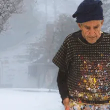 an older White Man with a paint-splattered sweater and knit cap stands in front of a snow-covered house.