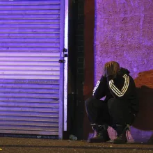 A Black Man in a track suit leans against a city strip wall with his head in his hands