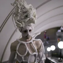 Young person, wearing white makeup, black contact lenses, white outfit made of tubes and a tall hand-made headdress, is taking an escalator in subway.