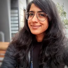 Woman of Indian descent with long black hair and glasses takes a selfie with a blurred background