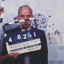 white male standing behind clapper board and in front of map labeled Riotsville with marked locations 