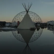 a white tipi sits in front of a steel geometric dome, both are reflected in a shallow pool at sunset.