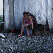 two female siblings sit on a plastic box and pet their black dog in front of a rural dilapidated barn.