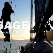 Jessie hangs from the ropes of her sailboat, peering into the sunset over the horizon.