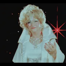 Ruth E. Norman dressed in white elaborate clothes beckons towards us with her hand out.