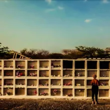 Sonia, a woman with dark skin, stands in her cemetery. She looks at a wall of graves. The text "People Like Us" is overlaid on top.