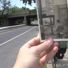 A hand holds an old photograph of a man with a bicycle at South Amboy Bridge. The photo lines up perfectly with South Amboy Bridge in the background. 