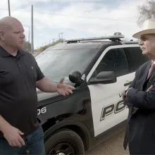 A man in a black shirt and a man in a suit and hat talk in front of a police cruiser.