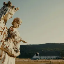 Large, white stone figure of Mary and baby Jesus in the foreground with barn and hills in the background. 