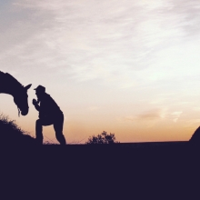 a man leans over to his horse, both silhouetted in the fading sunset.