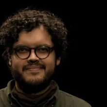 Manuel Acuna is pictured before a black background. He has curly dark brown hair, mustache and a beard. He is wearing black framed glasses and an olive green casual shirt.