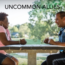 A Black woman sits across a park picnic table from a White Cop in uniform