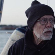 an older White man in glasses and a knit cap stands on the deck of a sailboat