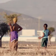 An Ethiopian farmer wearing a yellow headscarf and flowery dress and her daughter in ponytail wearing a blue T-shirt and green skirt stand in dry teff field looking into the distance against a hazy background of a massive industrial zone. 
