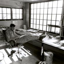 a black and white image of Wendell Berry reading a book in a chair with feet kicked up on table
