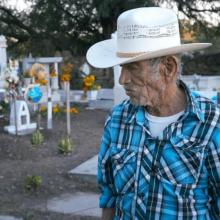 An elderly man in a white sombrero and blue checked shirt stands in a cemetery and directs his gaze downward. Behind him are graves decorated with flowers and balloons.