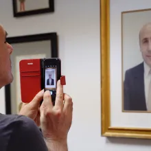 Bald white man takes a photo with an iPhone of a framed portrait of Alex Odeh. 