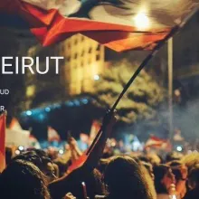 a protestor waves the red and white striped flag of Beirut over a sea of other protestors at night.