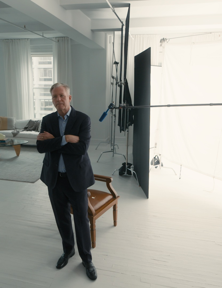 In a studio, Chris Hansen stands with his arms crossed, looking at a bearded white man in a white jacket.