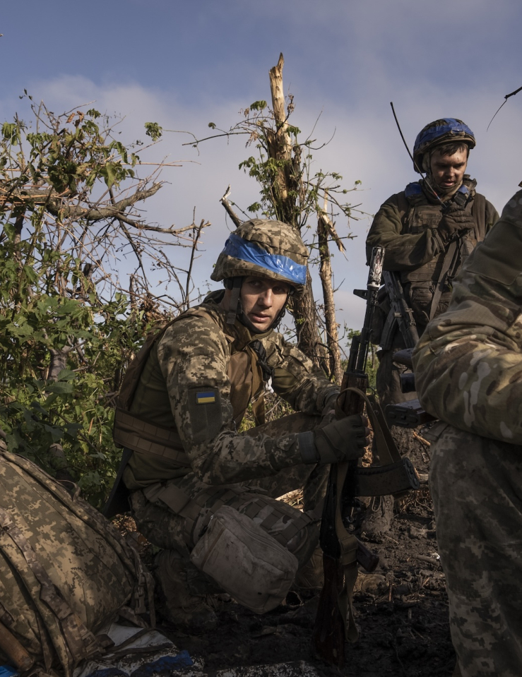 Three soldiers, with helmets and a small Ukrainian flag on their uniforms, crouching holding their guns outdoors