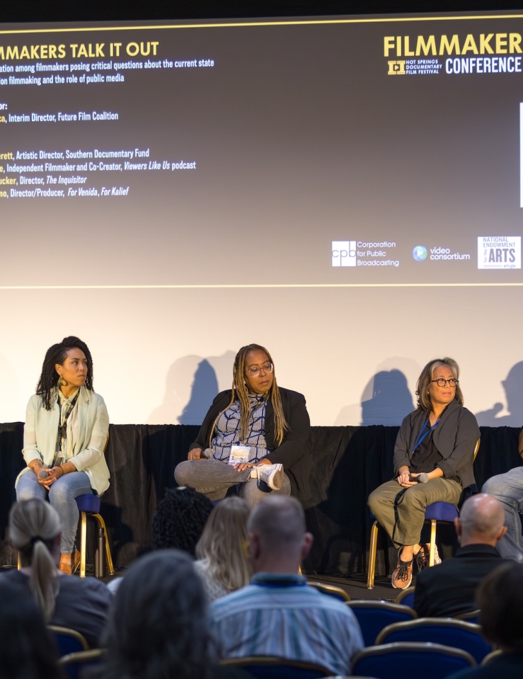 With a movie screen behind them that details the information for this Filmmaker Forum panel, four seated panelists with microphones face a crowd