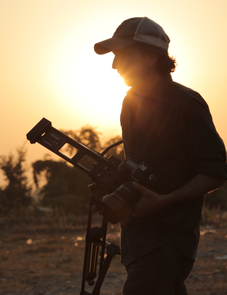 A man wearing a baseball cap and carrying a camera and tripod strolls through an arid landscape, illuminated by a sunset.