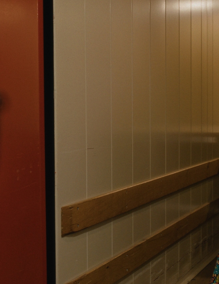 A camera on a tripod captures a portrait of a woman wearing a pink sweater, latex gloves, and a wooden indigenous mask in a nondescript room.