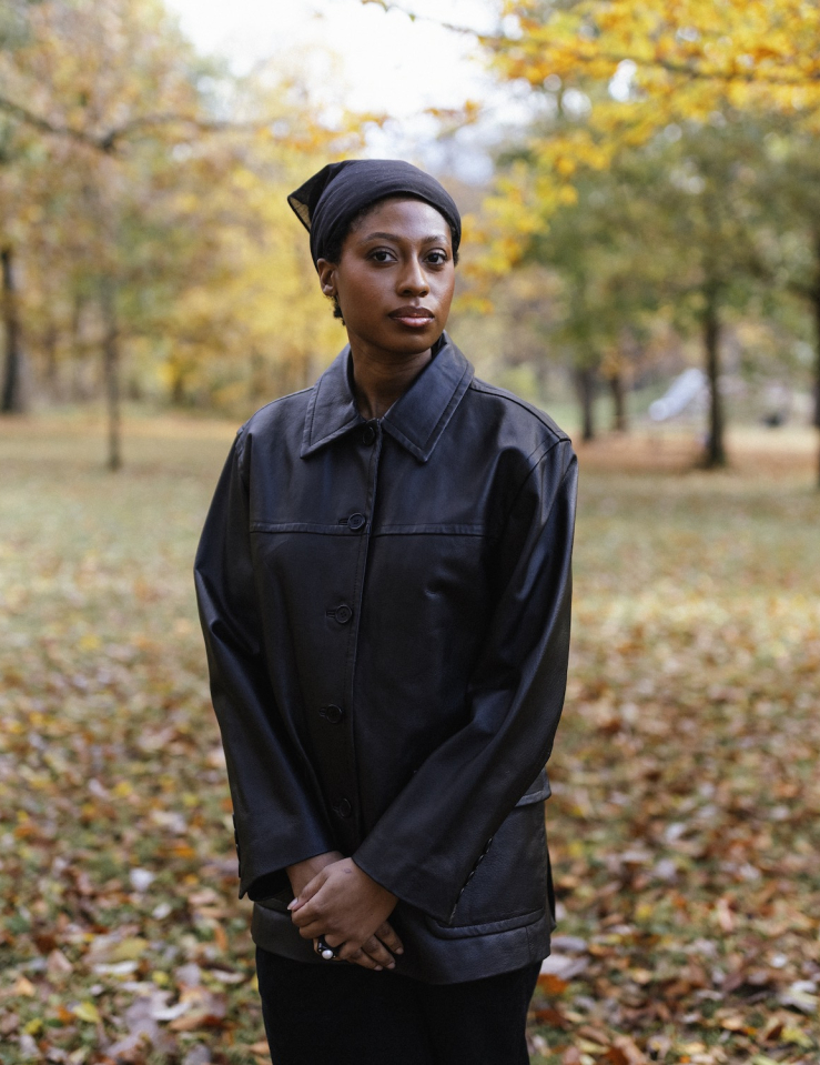 A Black woman in a black leather jacket stands with hands clasped in a field of autumnal leaves.