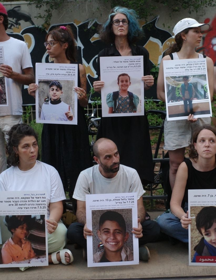 Two rows of people stand in silent vigil outside, holding large print-out posters of children's faces.