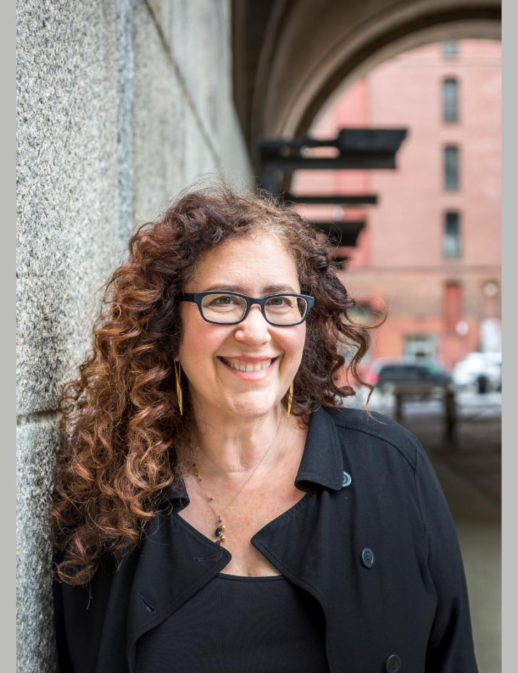 A vertical image of a smiling white woman with long curly brown hair, wearing glasses and a black shirt, leaning against a stone wall.