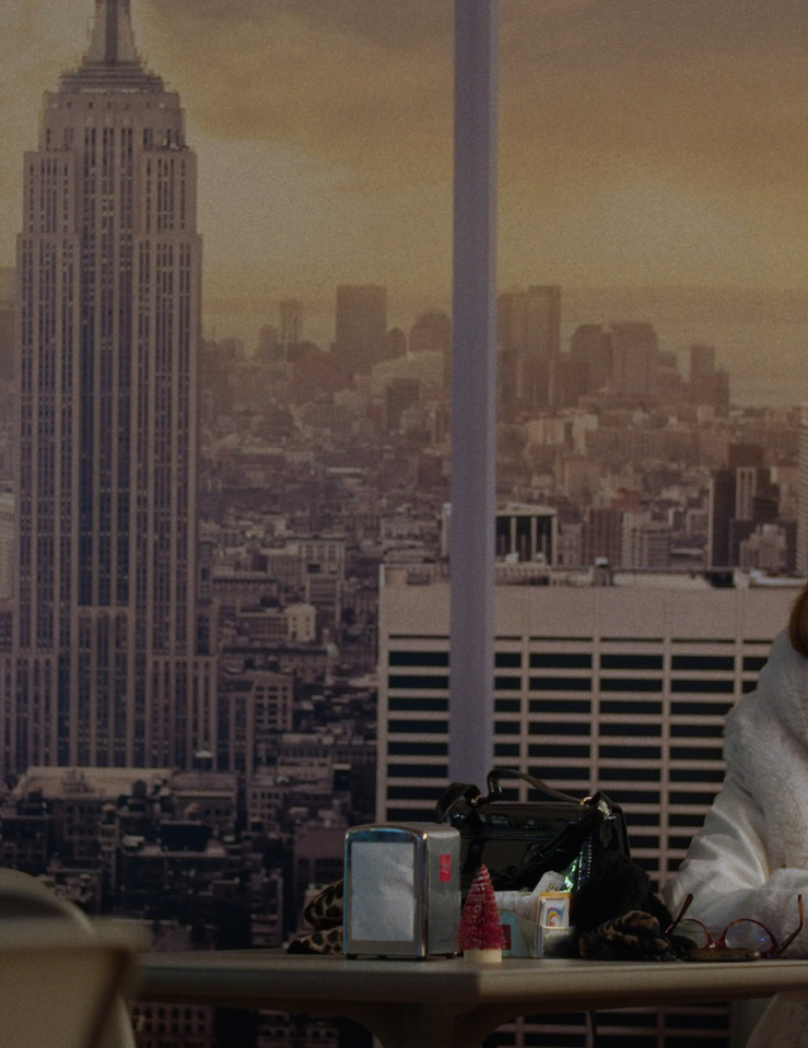 A woman with shoulder-length auburn hair wearing a white winter coat sits alone at a table with a blown-up print of the New York City skyline behind her