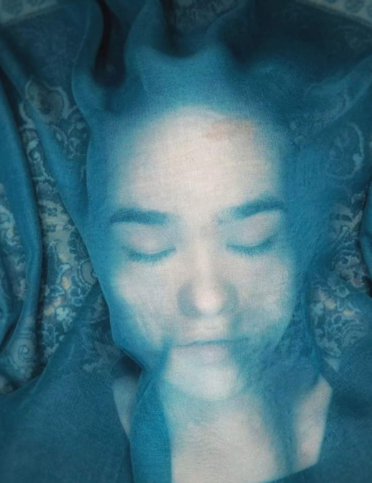 Under a blue cloth that is semi-transparent, a teenaged girl lays on the floor with her eyes closed.