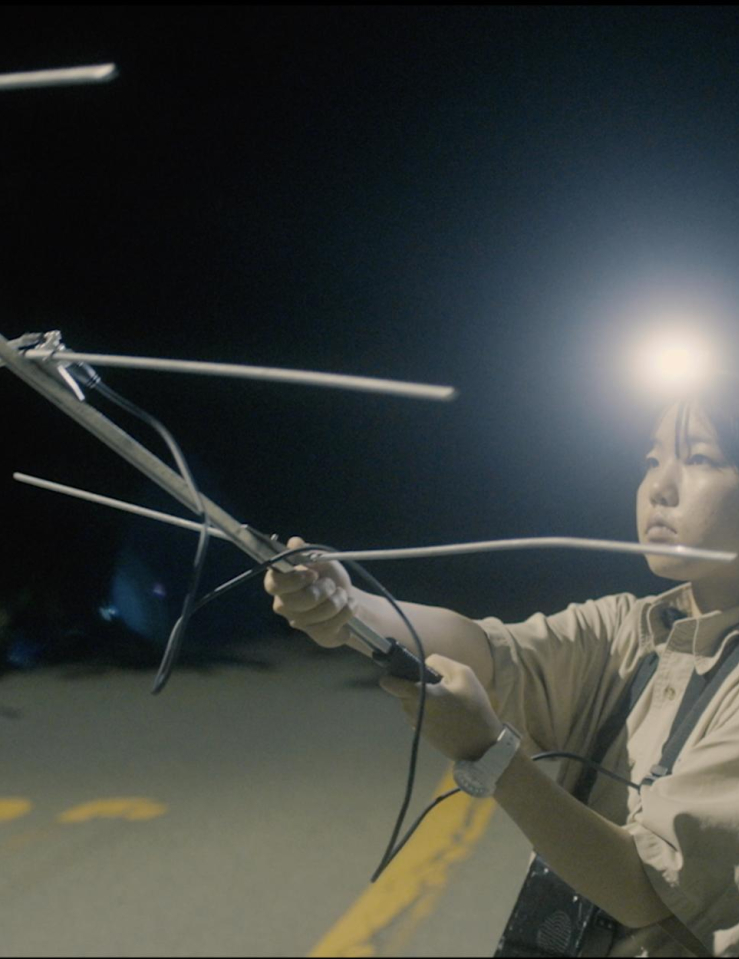 A young Asian woman stands in the middle of the street at night holding an antenna, lit only by the headlamp she sports on her head