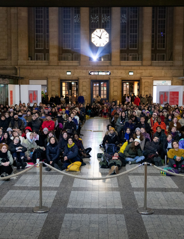 Hundreds of folks sit on the ground at Leipzig's Central Station, at a film screening; a projector can be seen behind them below a clock
