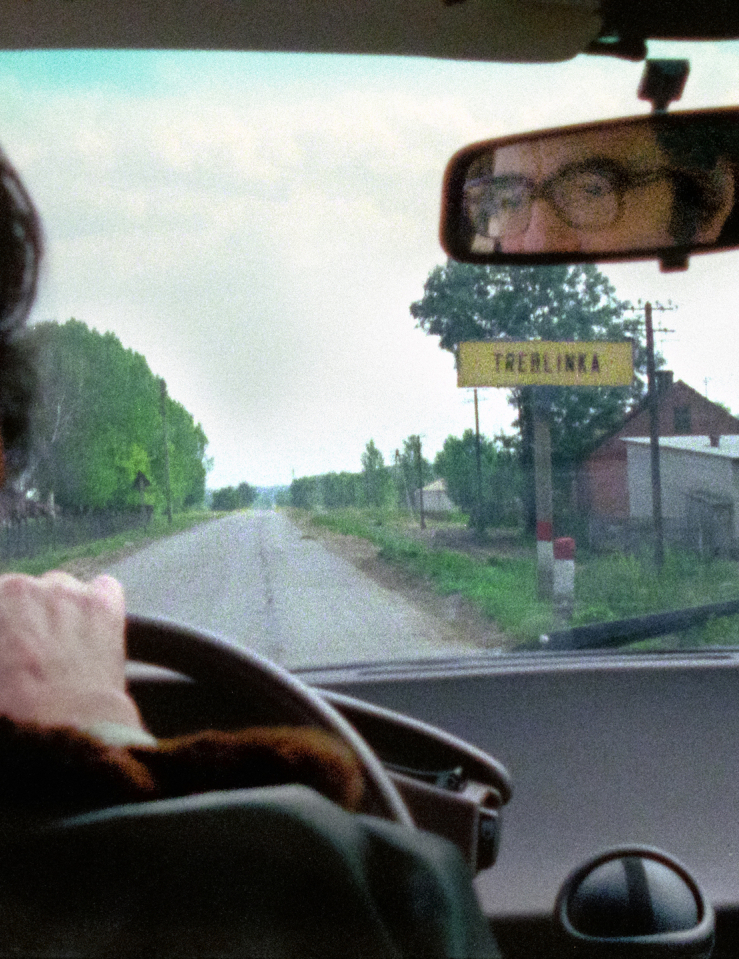 View from inside a car, a white man with dark hair and glasses can be seen driving through the rearview mirror as he passes a sign that reads Treblinka