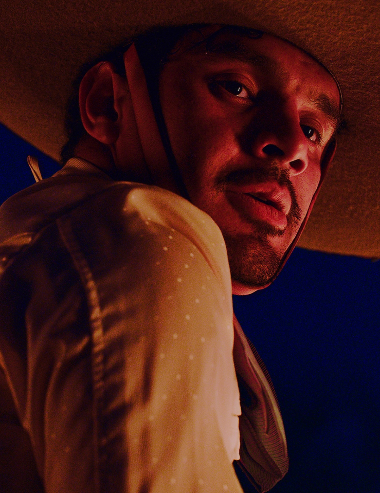 A brown-skinned young man wearing a mariachi suit and hat, lit by a red light looks down at the camera against a dark blue backdrop
