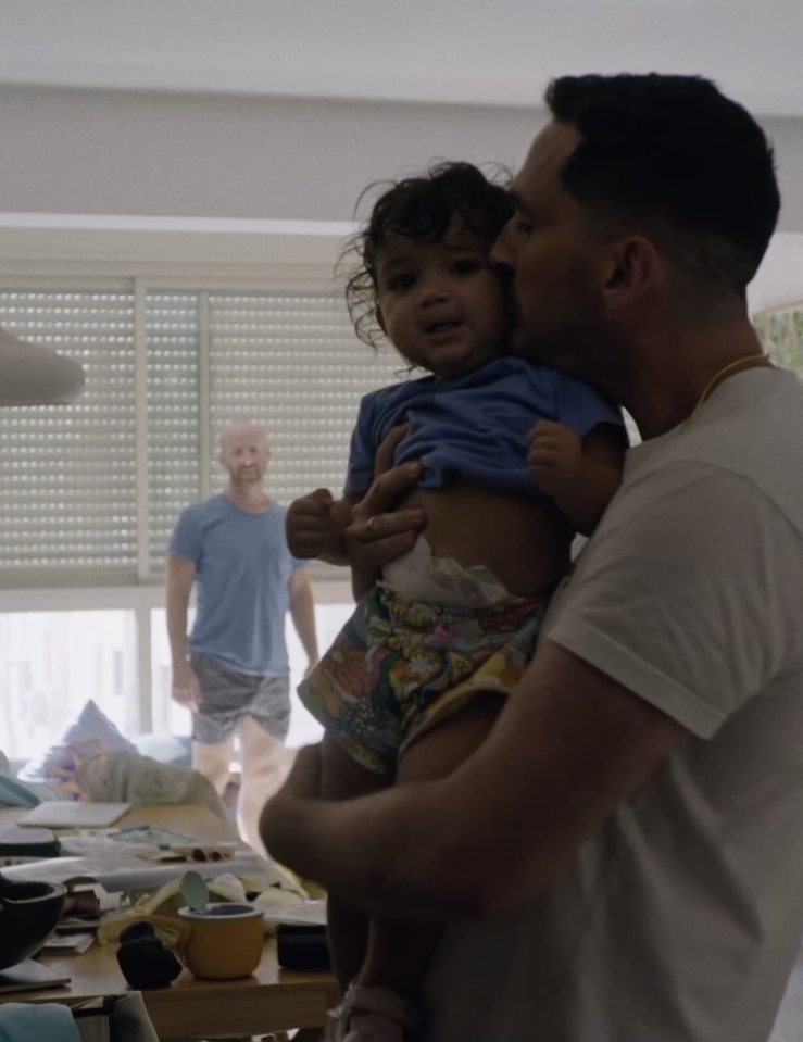 A young brown-skinned man hugs and kisses a young toddler girl amid a slightly messy living room while a balding white man in shirt and shorts looks at them in the background
