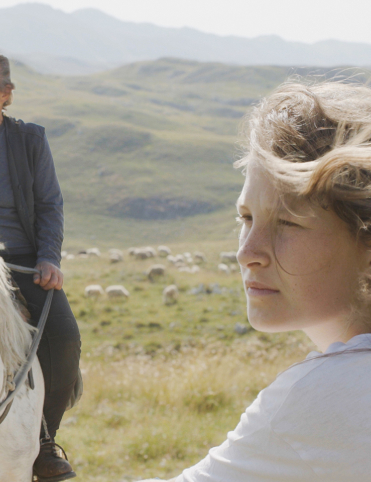 A young white blond girl in the mountains is seen in profile, while an older white woman with her grey hair pulled back rides a white horse