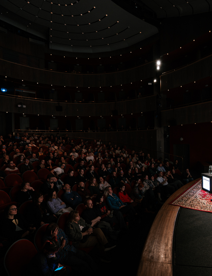 At a stage, two men sit in chairs as a rapt theater audience looks on