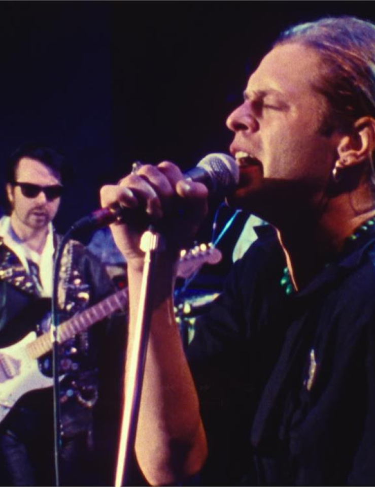 A man with a blond ponytail sings passionately into a microphone, in front of the rest of his rock band.
